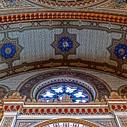 Ornate ceiling and stained glass window of a synagogue. Ornately tiled with blue, red, white, and brown. 