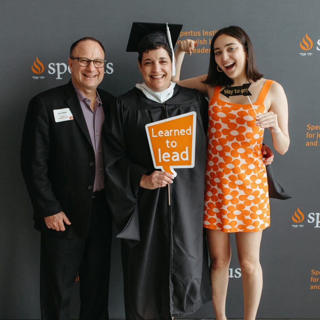 Three people stand in front of a wall with the Spertus logo printed on it. The woman in the center wears a black graduation cap and gown and holds an orange sign that says, "Learned to lead." The man on the left is smiling and wearing a black suit and glasses. The woman on the right wears an orange dress with flowers on it and holds a small black sign that says, "Way to go!"